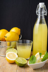 Top view of bottle with lemonade, glass, lemon wedges, lime, mint and whole lemons in metal bucket, on wooden table and black background, vertical