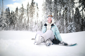 Fototapeta na wymiar Caucasian woman snowboarder sitting in snow near snowboard on beautiful snowy forest background in sunny day.