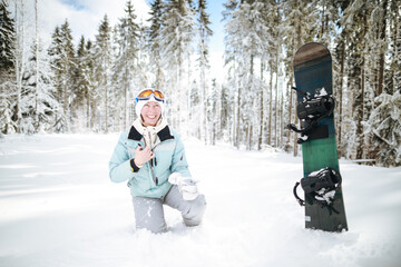Caucasian woman snowboarder sitting in snow near snowboard on beautiful snowy forest background in...