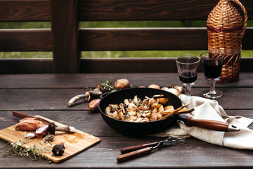 Roast forest mushrooms, potatoes, onions and seasonal herbs in a cast iron pan and ingredients on a wooden background outdoors.