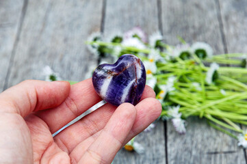 Amethyst heart-shaped gemstone in a hand, ox-eye daisy flowers in the background