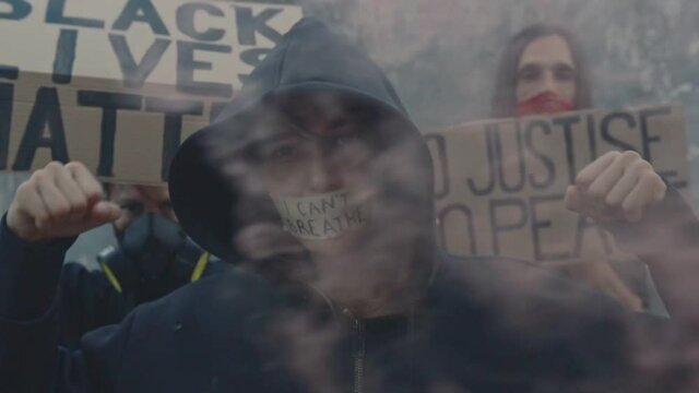 Young Caucasian woman in smoke with taped mouth and inscription I Can't Breathe on a peaceful protest manifestation against violence and racism, behind her protesters with placards Black Lives Matter