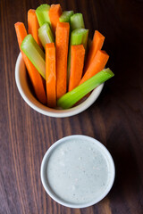 vertical close up isolated top view shot of a bowl of party snack in form of orange carrot and green cerely sticks with a white cup of blue cheese dipping sauce on a dark brown wooden table