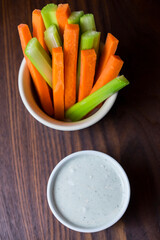 vertical close up isolated top view shot of a bowl of party snack in form of orange carrot and green cerely sticks with a white cup of blue cheese dipping sauce on a dark brown wooden table