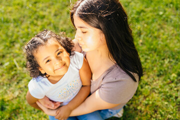 Top view portrait of ecuadorian single mother having funny conversation with her cute curly dark skinned hispanic daughter sits on green grass meadow in sunny summer day, outdoors.