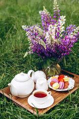 A cozy breakfast in the fresh air. A vase with lupine flowers, a teapot and a cup of coffee tea, a cake with macaroons on a wooden tray. Mother's Day or Women's Day. 