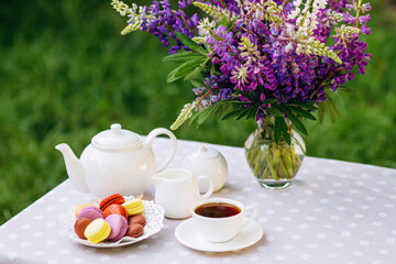 A cozy romantic breakfast in the fresh air. A vase with lupine flowers, a teapot and a cup of tea, a cake with macaroons on the table. Congratulations on Mother's Day or Women's. Soft selective focus.