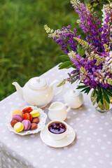 A cozy romantic breakfast in the fresh air. A vase with lupine flowers, a teapot and a cup of tea, a cake with macaroons on the table. Congratulations on Mother's Day or Women's. Soft selective focus.