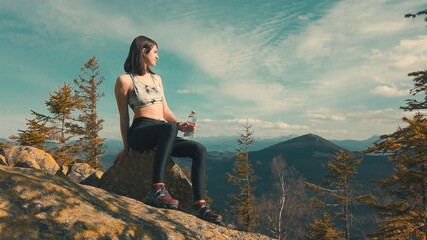 Portrait of beautiful athletic woman drinking clear water from a glass bottle while sitting on top of a mountain. Mountain peaks on a background. Girl enjoys the view around
