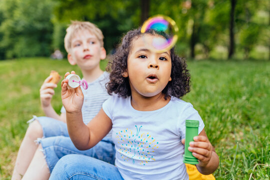 Happy multiethnic different age kids eating ice creams outdoor in summer. Cute positive hispanic girl enjoying blowing bulbs with her boy friend at sunny park. Summer, childhood, leisure concept