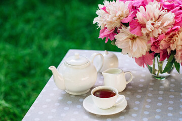 A cozy romantic breakfast in the open air. Peony flower vase, teapot and cup of tea on the table. Congratulations on mother's day or women's day. Soft selective focus.