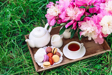 Cozy breakfast outdoors. Peony flower vase, teapot and cup of tea, macaroon cake on a wooden tray. Congratulations on mother's day or women's day. Soft selective focus.