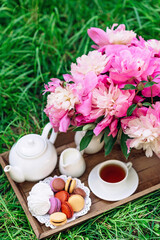 Cozy breakfast outdoors. Peony flower vase, teapot and cup of tea, macaroon cake on a wooden tray. Congratulations on mother's day or women's day. Soft selective focus.