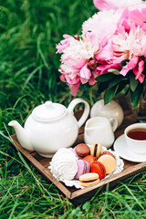 Cozy breakfast outdoors. Peony flower vase, teapot and cup of tea, macaroon cake on a wooden tray. Congratulations on mother's day or women's day. Soft selective focus.