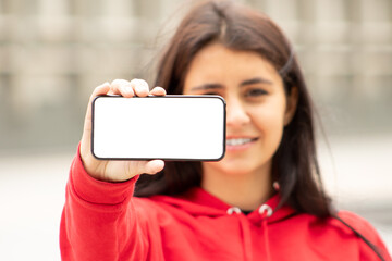 Front view portrait of a happy dark woman in red showing smartphone with blank screen on the street