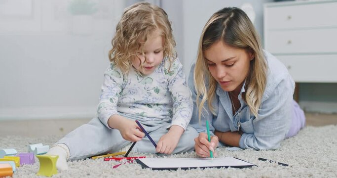 Middle plan of cute adorable kid girl holding color pencil learn drawing lying on warm floor with young mom daycare babysitter talking playing teaching helping little child daughter at home.