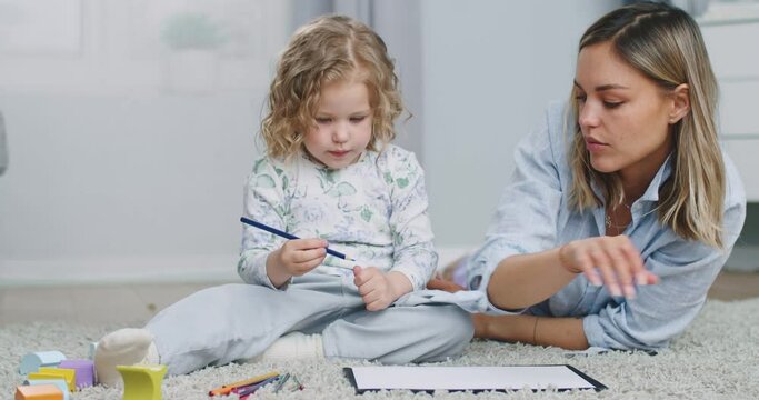 Middle plan of young caucasian mother helping her daughter to color the picture. Adult woman and little cute girl painting as laying at the flour at home. Creative family resting at weekends.