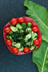 Vertical image.Summer salad.Top view of red plate full of cherry tomatoes, slices of cucumber, dill and horseradish leaves on the dark kitchen table