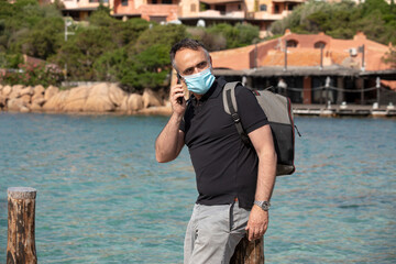 Uomo con mascherina facciale , maglia nera e zaino in spalla sta in vacanza  e sullo sfondo ha il mare e una località turistica 