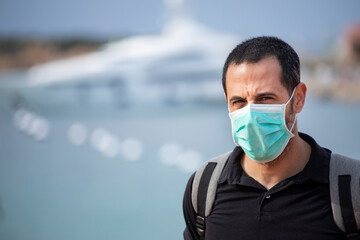 Uomo con mascherina facciale , maglia nera e zaino in spalla, è in vacanza  in piedi e sullo sfondo il mare con un grande yacht ormeggiato 