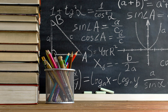 Books, pens, pencils and glasses on the table, against the background of a chalkboard with mathematicsformulas. Concept, teacher's day, knowledge day.