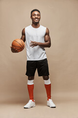 full length photo of a black athletic basketball player in the studio on a beige background wearing white T-shirt, black shorts, red long socks and white sneakers, he holds the ball and smiles