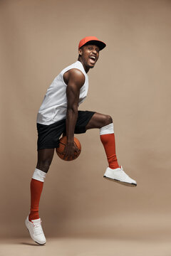 full-length photo of a black  athletic basketball player in the studio on a beige background, jumping high with a ball, wearing white T-shirt, black shorts, red long socks and a cap and white sneakers
