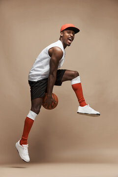 full-length photo of a black  athletic basketball player in the studio on a beige background, jumping high with a ball, wearing white T-shirt, black shorts, red long socks and a cap and white sneakers