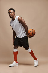 full length photo of a dark-skinned athletic basketball player in studio on a beige background posing with a ball wearing a white T-shirt, black shorts, red long socks and white sneakers