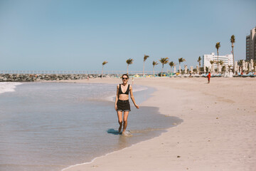 Young fit girl runs along the beach in a black swimsuit and shorts. She is smiling and happy.