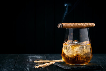 A glass of whiskey or cognac with ice cubes and a Smoking cigar lying on top, black wooden...