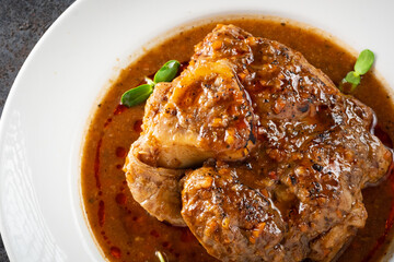 Stewed Osso Buco dish with tomato gravy on a white plate and a stone background, close up