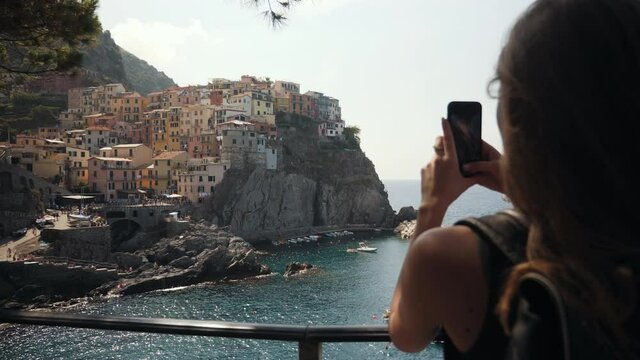 Young Woman Using Smartphone in the City Manarola Italy, Taking Pictures. SLOW MOTION 4K. Girl using cell phone photography app. Girl stands on the background of the city