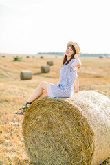 Naklejka na ściany i meble Young brunette Caucasian girl wearing straw hat and blue striped dress, sitting on a hay bale and posing at camera. Summer portrait of young woman in beautiful wheat harvesting field at sunset