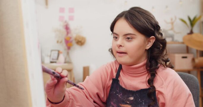 Front view of beautiful young lady smiling while painting picture in room. Sympathetic teenager girl with down syndrome sitting in front of molbert while having art therapy.