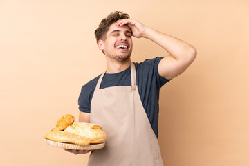 Male baker holding a table with several breads isolated on beige background laughing