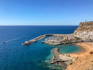 Fototapeta na wymiar View from the air on the coast and bay in the city of Mogan on the Canary Islands where the bay harbor and people walking along the waterfront inside city