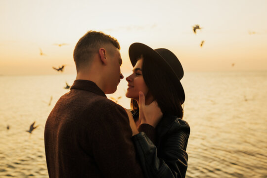Portrait of happy couple in love dressed in stylish classical clothes are hugging sensually during a beautiful summer sunset near the sea. Close-up photo. Flying seagulls. Copy space.