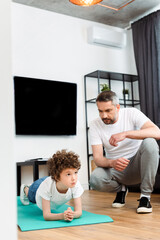 bearded father looking at curly son working out on fitness mat