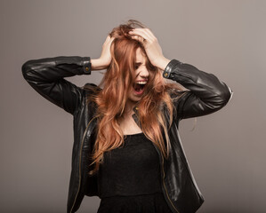 Fototapeta premium Tired from work blond hair female model with agressive emotions crying with wide opened mouth and holding head. Style clothing woman in black leather jacket on grey studio background. Closeup