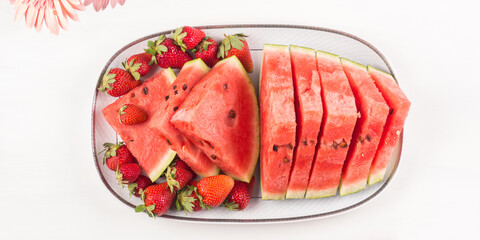 Watermelon slices with strawberries on ceramic tray on white wooden table with pink flowers. Fresh summer snack