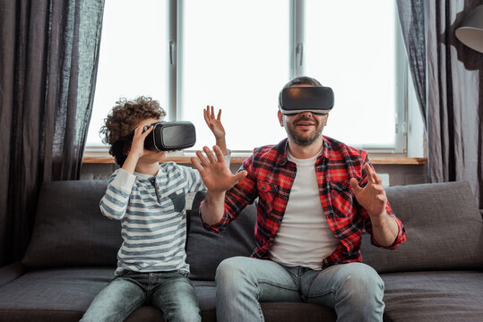 bearded father and curly son in virtual reality headsets gesturing in living room