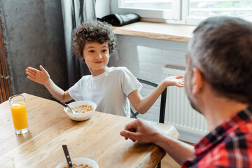 selective focus of curly boy gesturing and looking at father near bowls with corn flakes and glass of orange juice