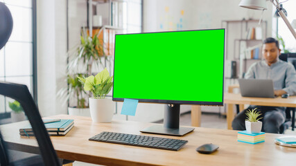 Mock-up Screen Desktop Computer Standing on the Desk in the Modern Creative Office. In the Background Bright Studio Apartment where Diverse Team of Young Professionals Work on Computers.