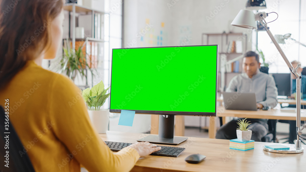 Poster over the shoulder: creative young woman sitting at her desk using desktop computer with mock-up gree