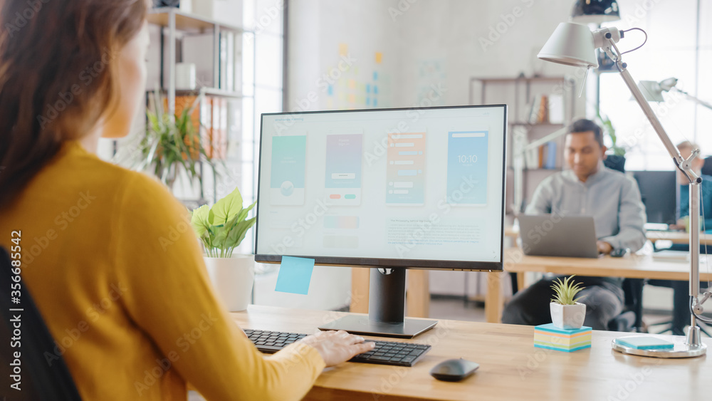 Poster Over the Shoulder: Female Mobile Software Developer Sitting at Her Desk Using Desktop Computer with Screen Showing Smartphone Application UI / UX. Office With Diverse Team of Professionals Working.