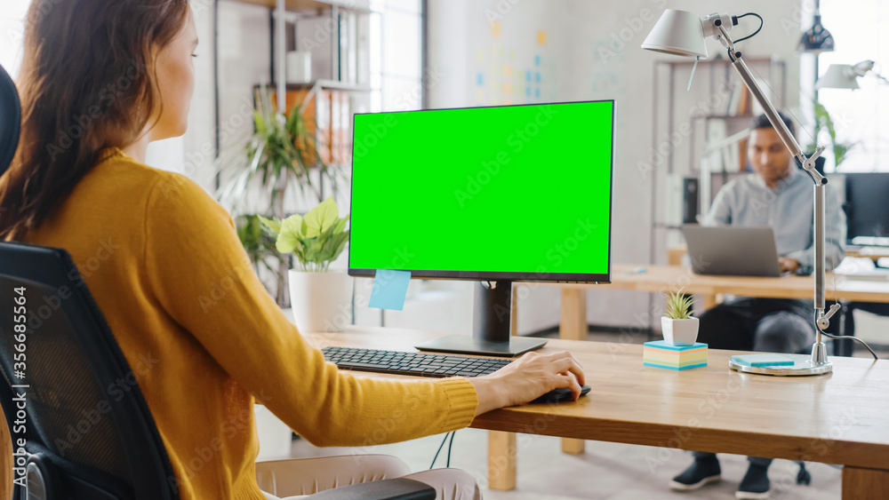 Poster over the shoulder: creative young woman sitting at her desk using desktop computer with mock-up gree