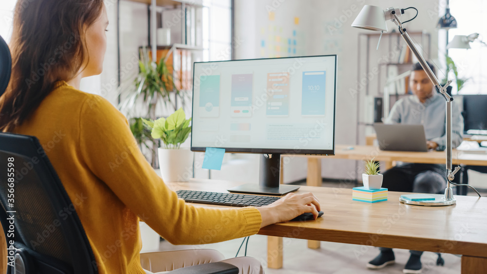 Poster Over the Shoulder: Female Mobile Software Developer Sitting at Her Desk Using Desktop Computer with Screen Showing Smartphone Application UI / UX. Office With Diverse Team of Professionals Working.