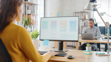 Over the Shoulder: Female Mobile Software Developer Sitting at Her Desk Using Desktop Computer with Screen Showing Smartphone Application UI / UX. Office With Diverse Team of Professionals Working.