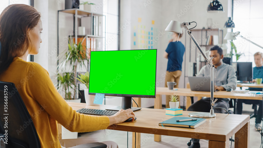 Poster over the shoulder: creative young woman sitting at her desk using desktop computer with mock-up gree